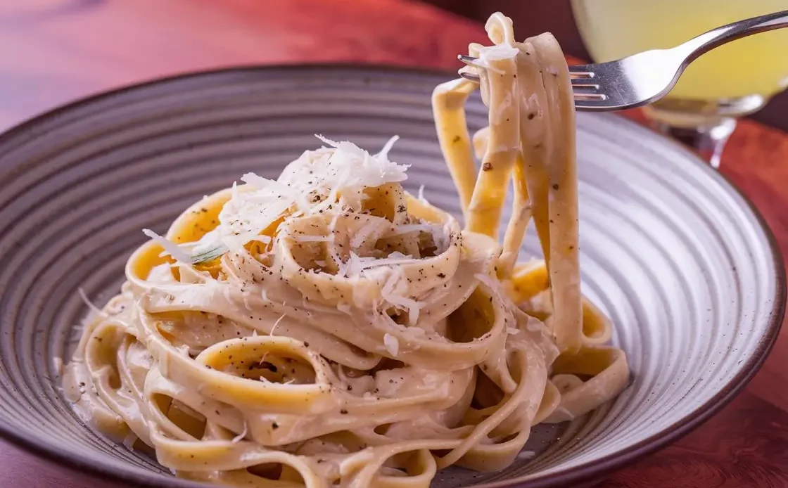 A gourmet plate of Black Pepper Fettuccine, freshly cooked, served on a ceramic plate. The pasta is coated in a creamy Pecorino Romano sauce, with visible flecks of freshly ground black pepper. The dish is garnished with extra grated cheese and cracked pepper.